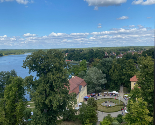 Noch einmal der Blick hinunter auf Schlosspark und See – eingerahmt von viel Grün und einem erstaunlich weiten Himmel. Genau für solche Aussichten steigt man 146 Stufen hinauf.